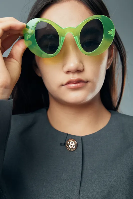 Young woman with long black hair wearing large green sunglasses and a dark gray top with button posing isolated on gray background for fashion and style portrait.