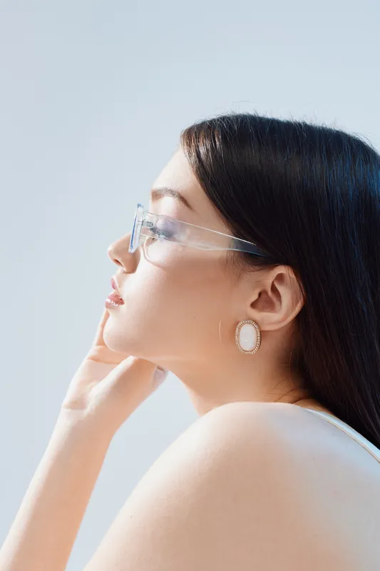 Profile of a young woman with long dark hair and clear glasses wearing elegant earrings. Soft natural light highlights her smooth skin and thoughtful expression. Beauty fashion concept.