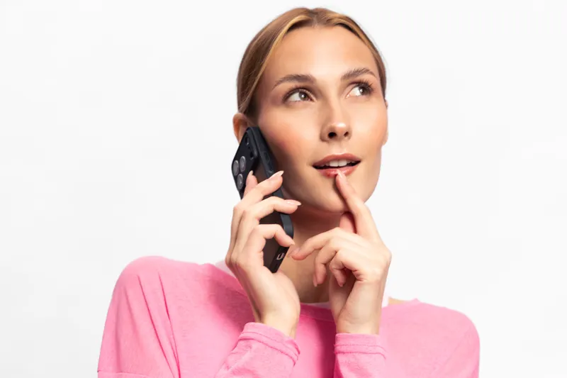 Young woman in pink sweater talking on phone with thoughtful expression isolated on white background. Concept of communication, technology, and casual lifestyle.
