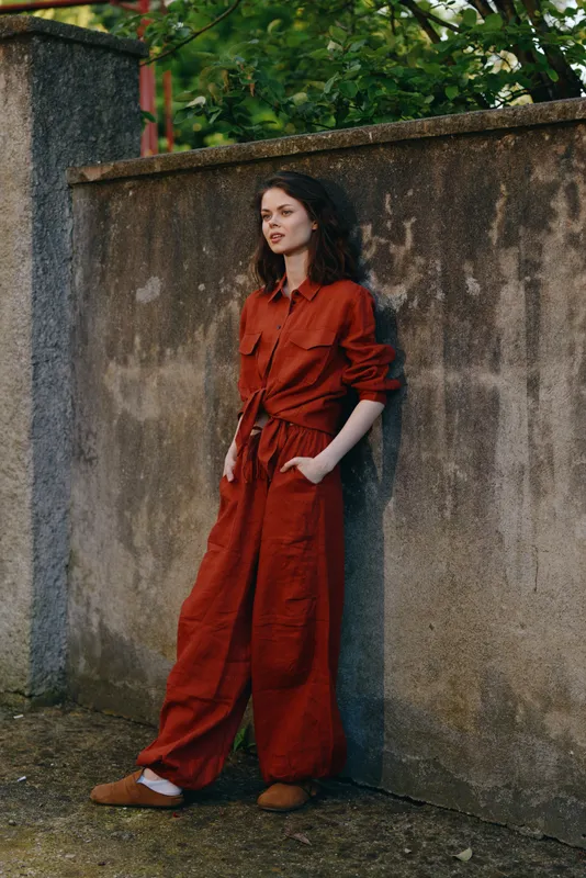 Woman in a red jumpsuit leans against a weathered wall, outdoor lifestyle portrait, relaxed pose, soft natural light, stylish casual fashion moment, captured with authentic mood and contemporary