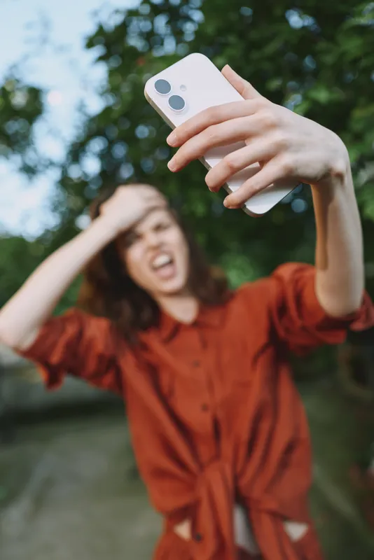 Young woman outdoors takes a selfie, smiling while raising a phone, wearing an orange dress against a leafy natural background