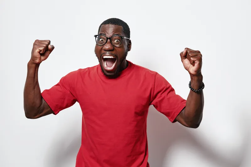 African American man with dark skin wearing glasses and red t shirt celebrating with raised fists and open mouth for excitement and success on white background.
