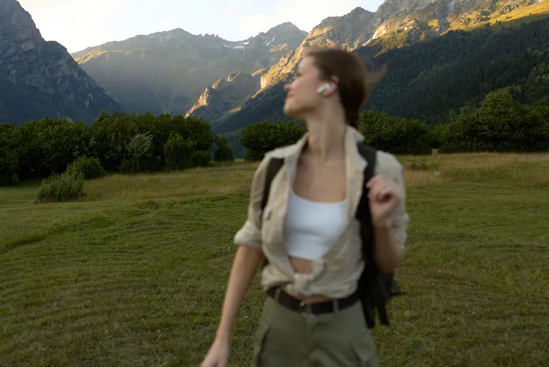 A woman in light outdoor attire walks through a grassy meadow with distant mountains, carrying a backpack. Concept for solo travel, nature exploration, and casual lifestyle.