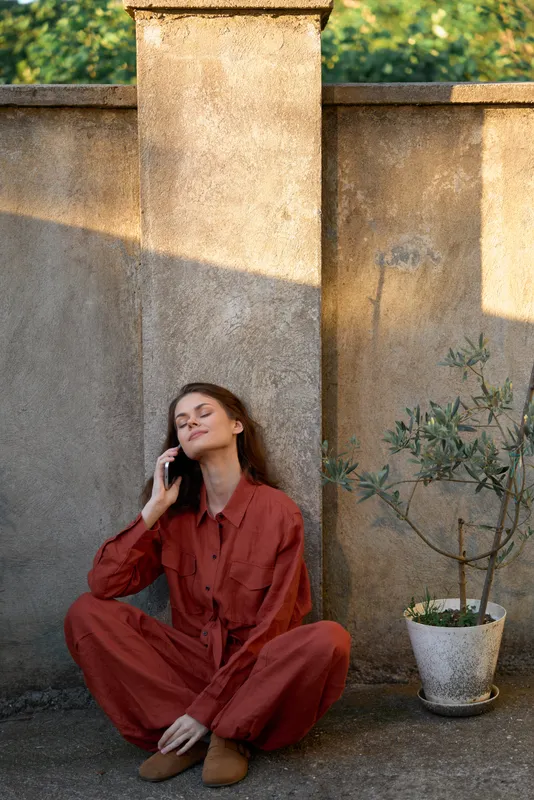 Relaxed woman in a rust jumpsuit sits against a sunlit wall, talking on phone, enjoying a calm outdoor moment in a courtyard setting