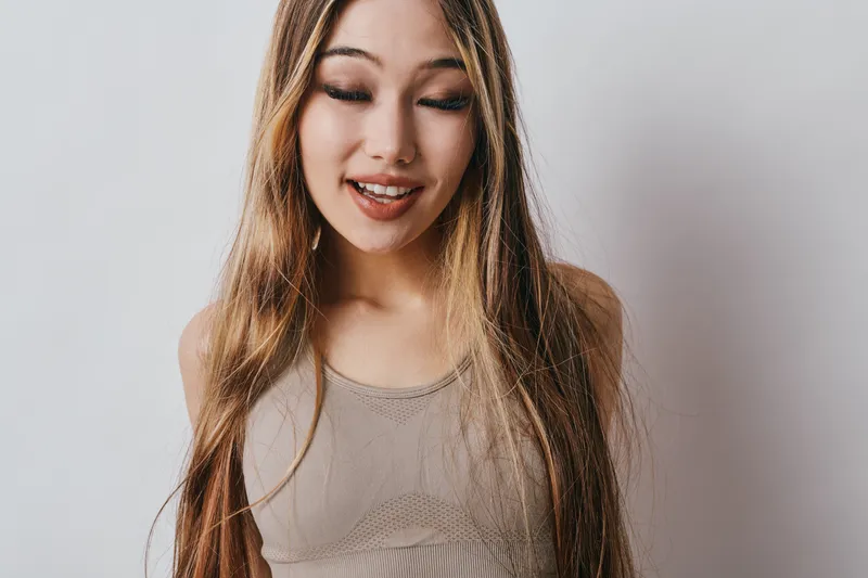 Young woman with long hair and closed eyes smiling naturally on white background. Casual style, relaxed mood, friendly female portrait with soft lighting.
