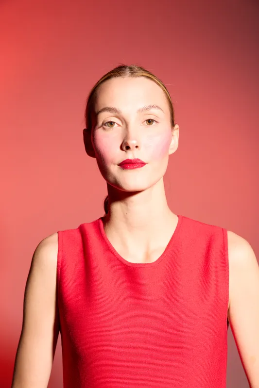 Young woman with fair skin and red lipstick wears sleeveless red dress against red background. Studio portrait with strong lighting highlights confident expression and fashion style.