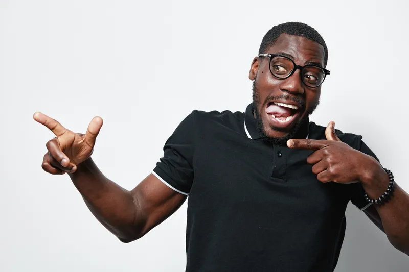 African American man with dark skin and glasses wearing black polo shirt pointing and smiling with excitement on white background. Casual, happy, confident expression.