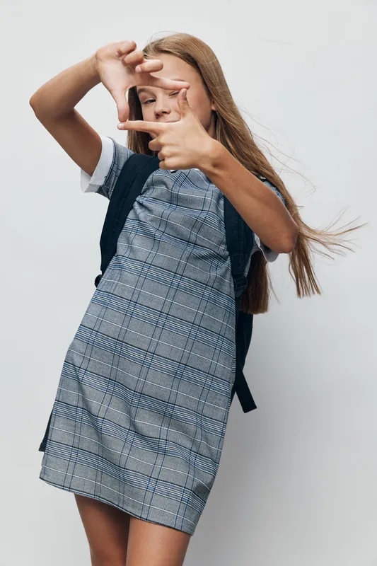 Girl with backpack posing with fingers frame in front of face, wearing plaid dress and white shirt against plain background, long hair and casual style for school or youth concept.