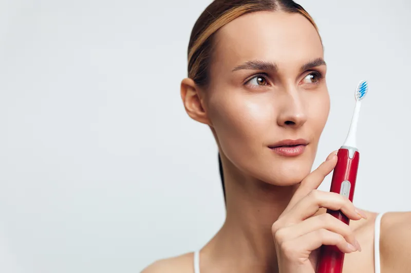 Smiling woman holding a red electric toothbrush. Close-up on a light background. Concept of personal care, skincare, and dental hygiene. Soft studio lighting creates a calm mood.