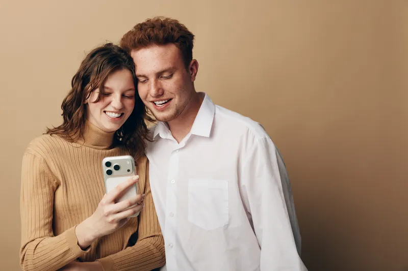 Smiling young couple looking at smartphone screen together, standing close on beige background. Casual clothing, happy mood, technology use, communication, and relationship concept.