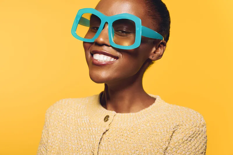 Smiling black woman with dark skin wearing large turquoise glasses and yellow sweater poses against bright yellow background. Concept of fashion, joy, and vibrant style.