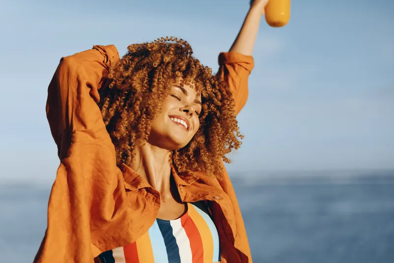 Young woman with curly hair raises a bottle outdoors, smiling in the sun by the sea, wearing a striped top and an orange jacket, joyful and carefree, with wind in her hair and a bright blue sky