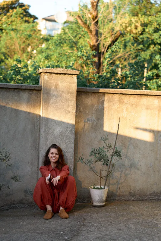 A young woman in a red jumpsuit sits on the ground beside a weathered wall, offering a relaxed, contemplative vibe as sunlight spills across the scene
