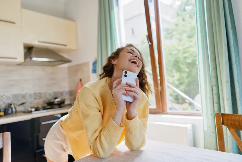 Cheerful woman at home, indoors, holding a smartphone and smiling, cozy kitchen setting, natural daylight, relaxed posture, candid lifestyle moment, soft colors, welcoming atmosphere