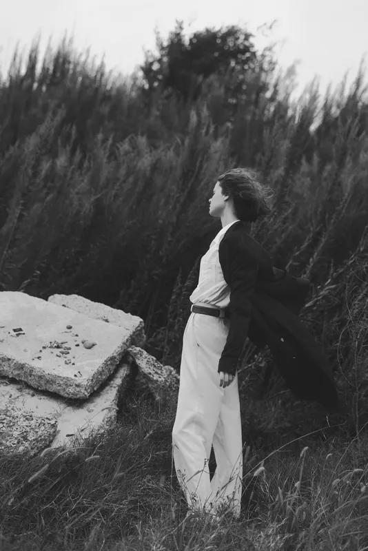 Woman standing in a windy field with a flowing coat, black and white atmosphere, cinematic mood, solitude and exploration, natural landscape, fashion influence, dramatic composition
