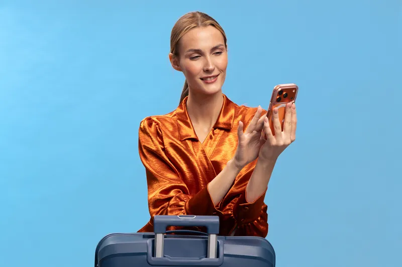 Young woman in orange satin blouse holds smartphone and smiles, standing behind blue suitcase on light blue background. Concept for travel, communication, and lifestyle.