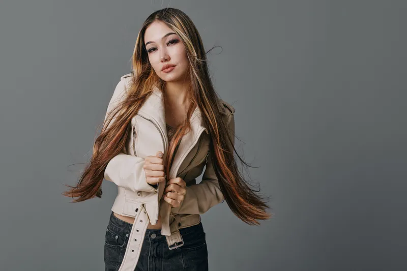 Young woman with long hair wearing beige jacket and black pants posing on gray background for fashion portrait and style concept indoors.