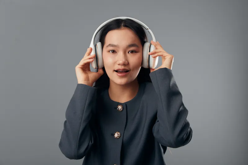 Young woman with headphones listening to music or podcast on neutral background. Portrait of female with hands holding wireless headset for enjoyment or communication in studio.