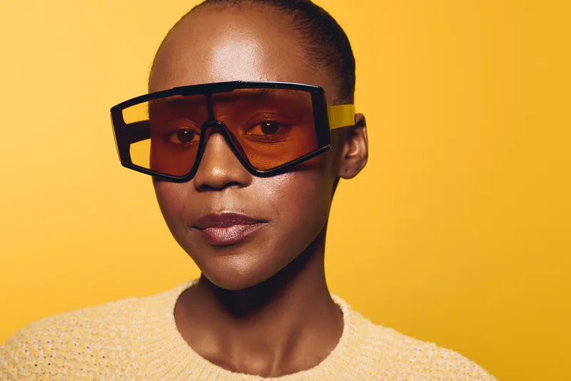Close-up portrait of a confident black woman with dark skin wearing oversized futuristic sunglasses and a yellow knitted sweater on a bright yellow background. Fashion concept.