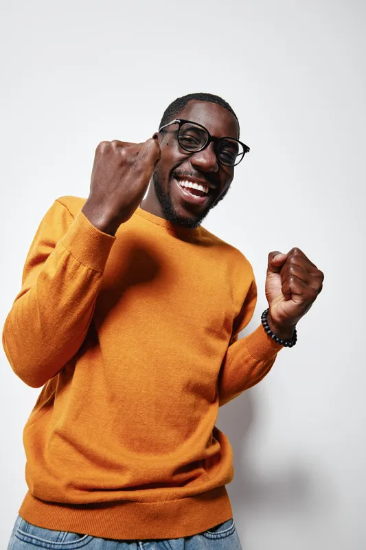 African man with dark skin wearing glasses and orange sweater smiling and celebrating success with raised fists against white background in studio portrait.