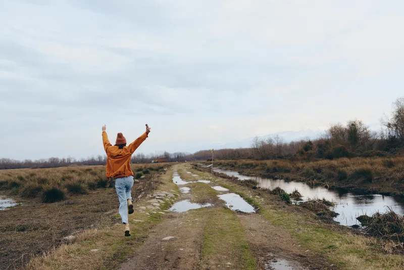 Woman outdoors lifestyle walking on river path, jump with arms raised, cheerful winter mood, jeans and sweater, cloudy sky, muddy puddles, shrubs and bare trees, calm nature, peaceful