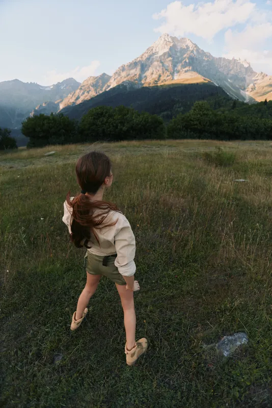 Cheerful girl running across a grassy field toward distant mountains, sunlit hair, carefree moment, natural colors, open landscape
