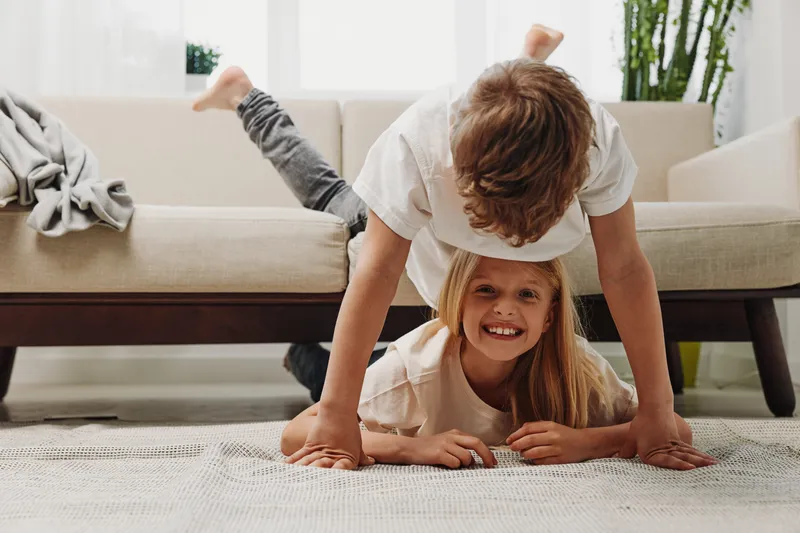 Happy children playing indoors on carpet, boy balancing on hands over smiling girl lying on floor near sofa in bright living room with plants. Concept of childhood fun and family.