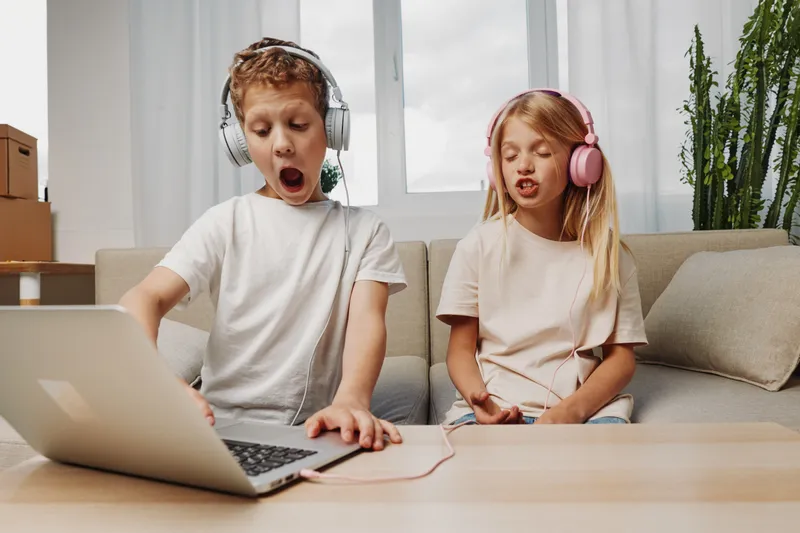 Two children with headphones enjoy music on laptop, expressing emotions while sitting on couch in bright living room.