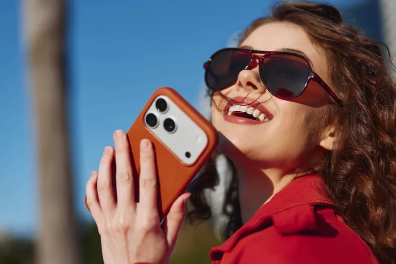 Young woman with curly hair wearing red sunglasses and a red jacket smiles joyfully while holding a smartphone outdoors on a sunny day.