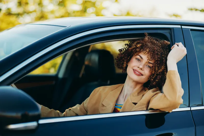 Vibrant portrait of a woman inside a car outdoors, sunlight on curly hair, relaxed smile, retro color vibes, casual fashion, candid moment in warm natural scenery