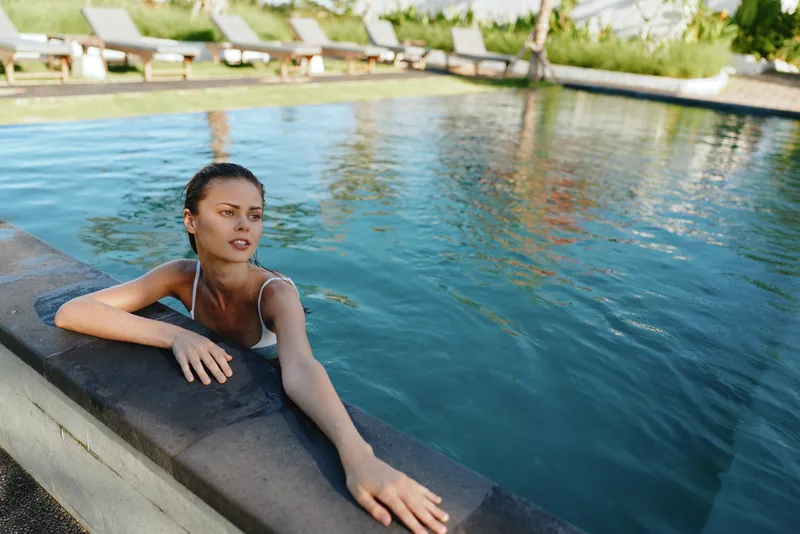 Woman leans on pool edge, hands resting in cool water. Outdoor poolside scene conveys relaxation, wellness, and leisure with warm sunlight, clear blue water, and a calm, inviting mood for lifestyle