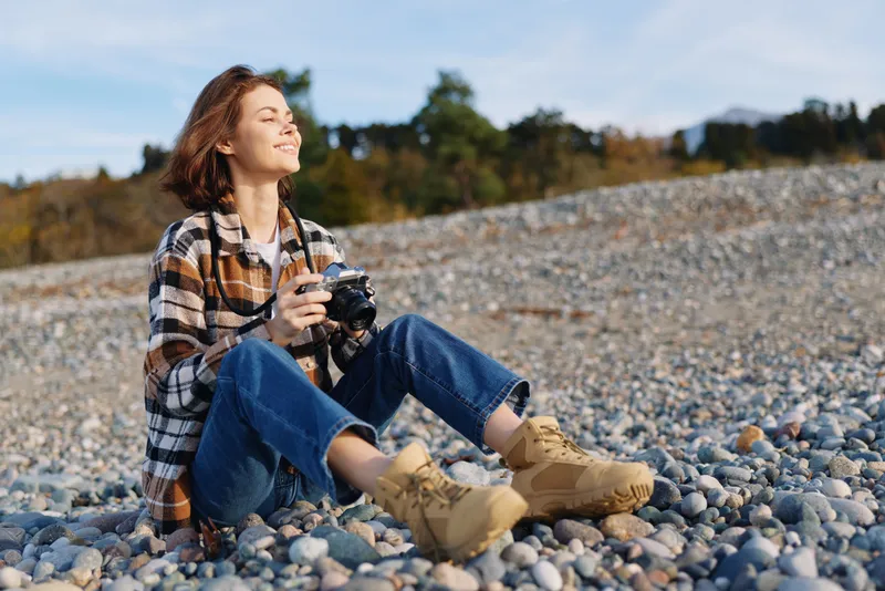 Outdoor woman photographer sits on a pebble beach, holding a camera and enjoying bright sun, wearing casual flannel shirt, jeans and boots, capturing simple nature moments.