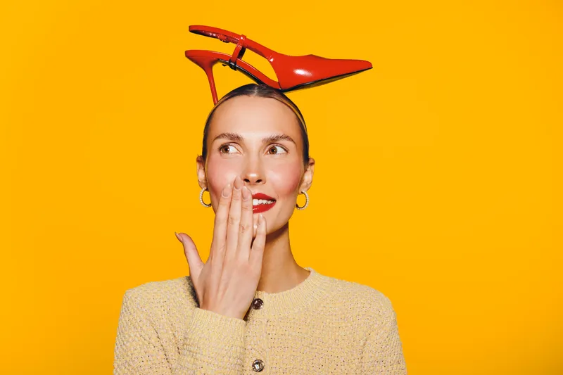 Young woman with red lipstick and earrings holds red high heel shoes on her head, smiling and covering mouth on bright yellow background. Concept of fashion, fun, and creativity.