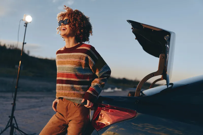 Lifestyle woman with curly hair stands by car outdoors, open trunk visible, warm sunset sky and sea background, casual sweater, relaxed travel preparation, candid moment