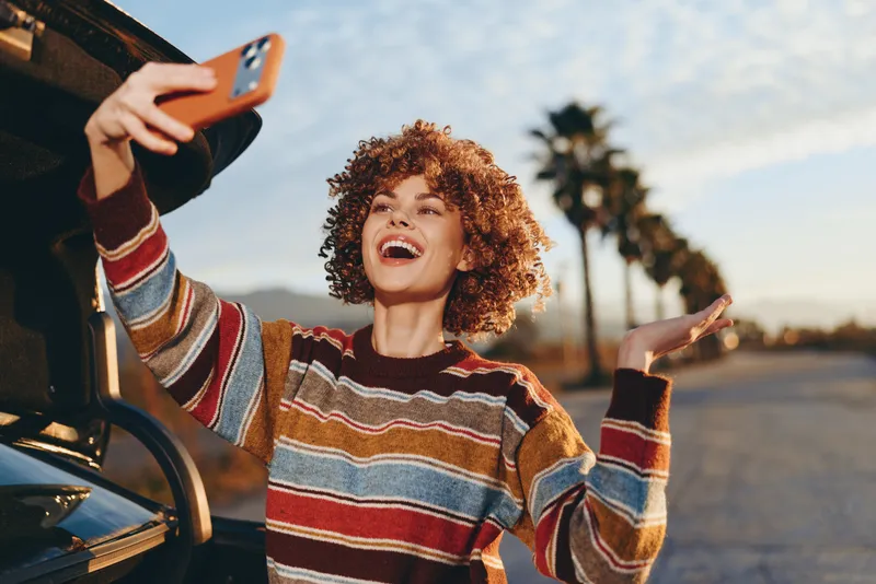 Woman with curly hair laughing for a selfie using a smartphone from the car, outdoors in daylight on a sunny road, casual lifestyle, warm cheerful mood, travel adventure.
