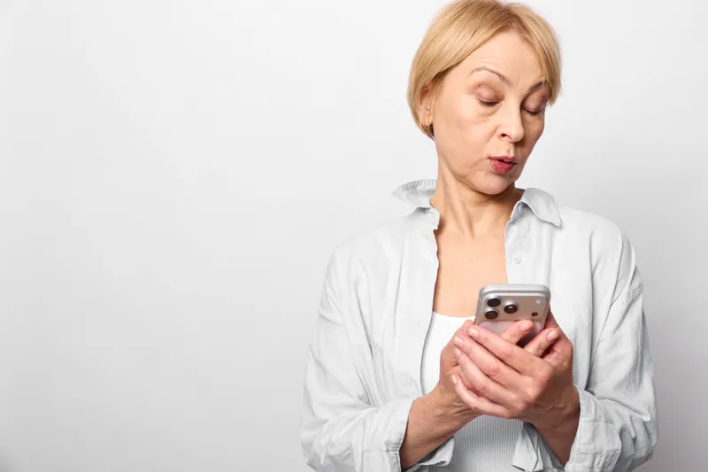 Woman smartphone technology communication portrait studio white mature texting indoors, blond short hair, casual shirt, holding a smartphone while looking down, calm concentrated face, studio light