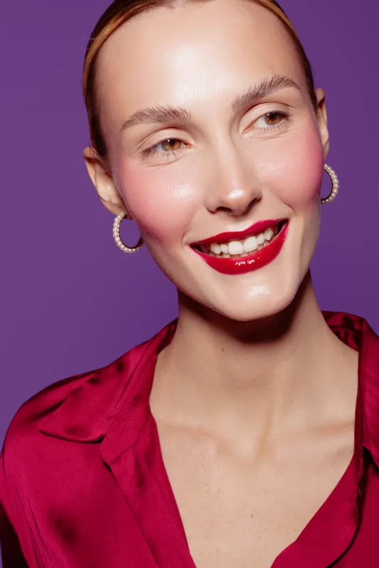 Smiling woman with red lipstick and hoop earrings wears a red satin blouse against purple background. Close-up portrait with natural skin and joyful expression. Beauty and fashion concept.