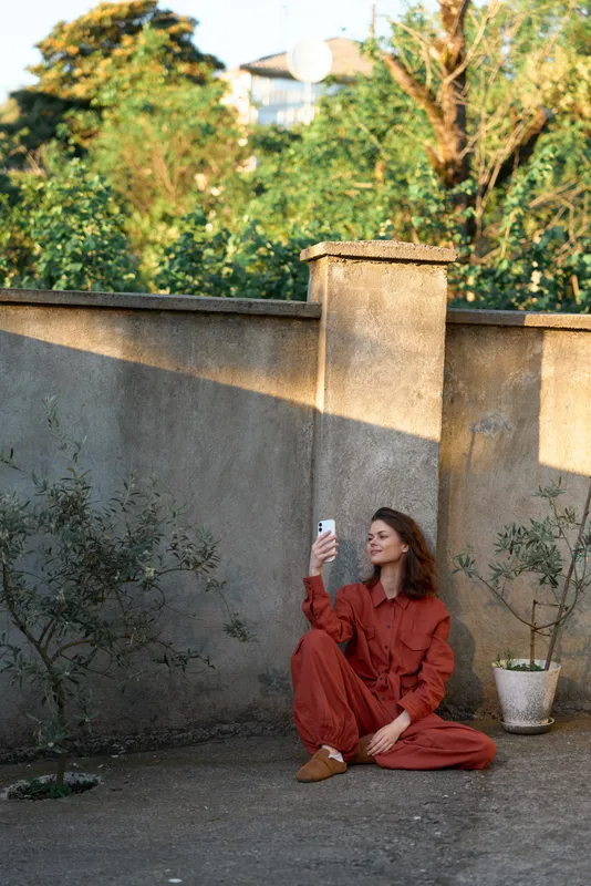 A woman in a red jumpsuit sits on a courtyard ground, taking a selfie with a smartphone. Bright natural light highlights casual street style and a calm, reflective mood