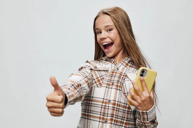 Young woman with long hair holding smartphone and showing thumbs up with happy smile isolated on grey background. Female expressing positivity and approval in casual shirt.