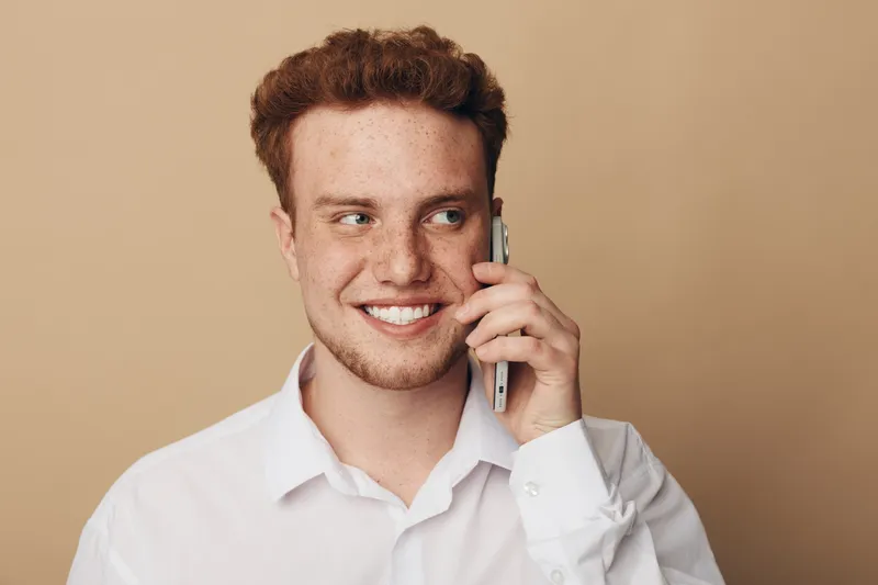 Smiling young man with curly hair wearing white shirt talks on smartphone. Close-up portrait on beige background. Concept of communication, technology, and positive emotions.