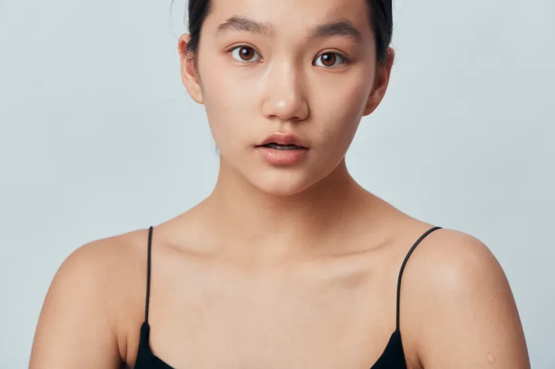 Portrait of young woman with natural skin and dark hair, wearing black spaghetti strap top, looking at camera with neutral expression on plain light background in studio.