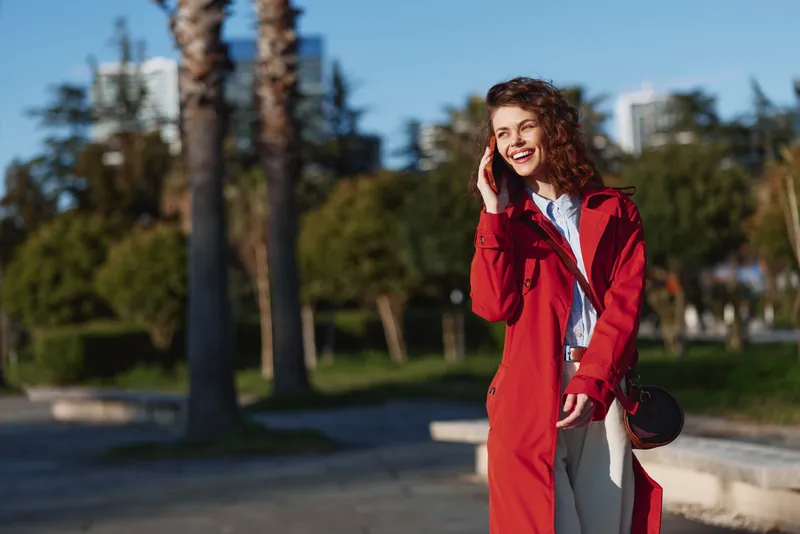 A smiling woman in a red coat stands outdoors on a sunny day, posing in a park setting, showcasing casual street fashion with an urban background.