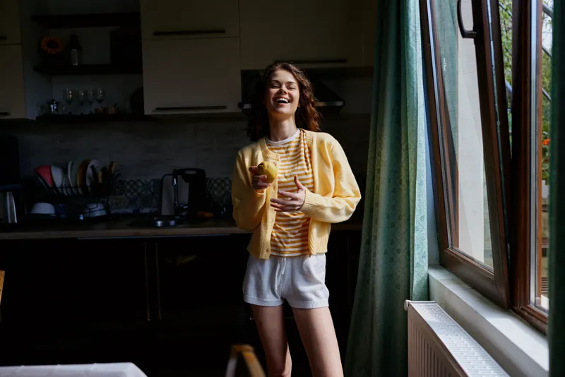 A cheerful woman stands in a sunlit kitchen, wearing a yellow cardigan and striped top, laughing while holding a glass. A genuine, everyday home moment about joy and comfort.