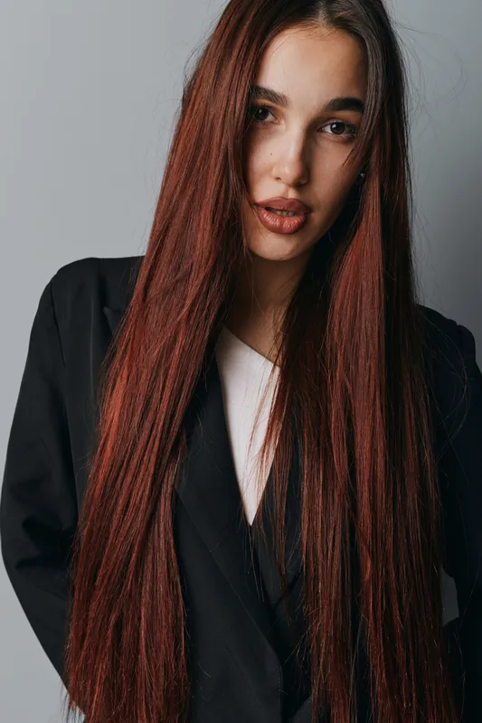 Young woman with long straight red hair wearing black blazer and white top posing against gray background in studio. Beauty, fashion and portrait with natural makeup and confident expression.