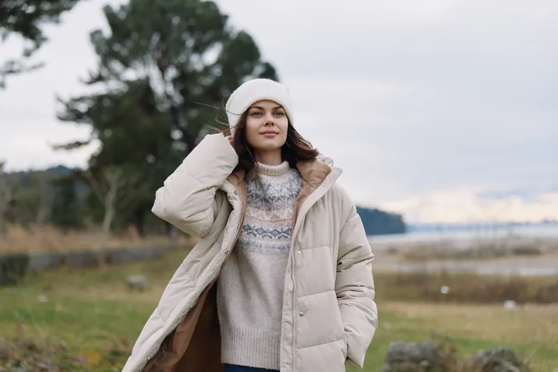 Young woman in a warm beige winter coat stands outdoors by a lakeside, wearing a knit sweater and beanie in a breezy rural landscape today.