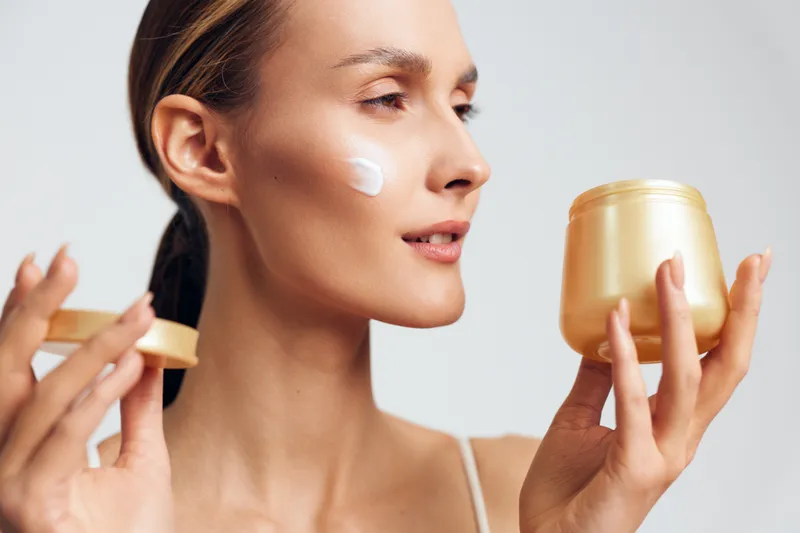 Young woman applying face cream on cheek with soft texture holding jar in hand against white background. Concept of skincare, beauty, wellness, and daily self-care routine.