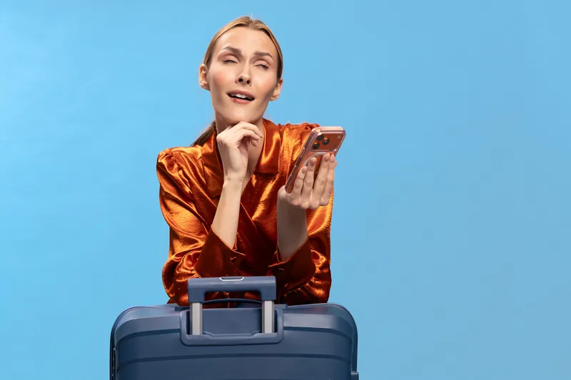 Thoughtful woman in shiny orange blouse holds smartphone and leans on blue suitcase against blue background. Concept for travel planning, decision making, and communication.