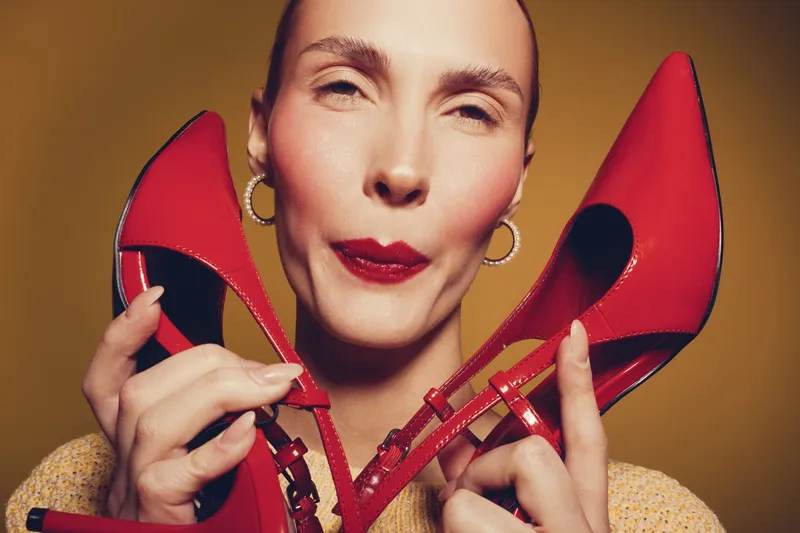 Close-up of a woman holding red high heels near her face with a playful expression. Studio portrait with warm lighting and yellow background. Concept of fashion and style.