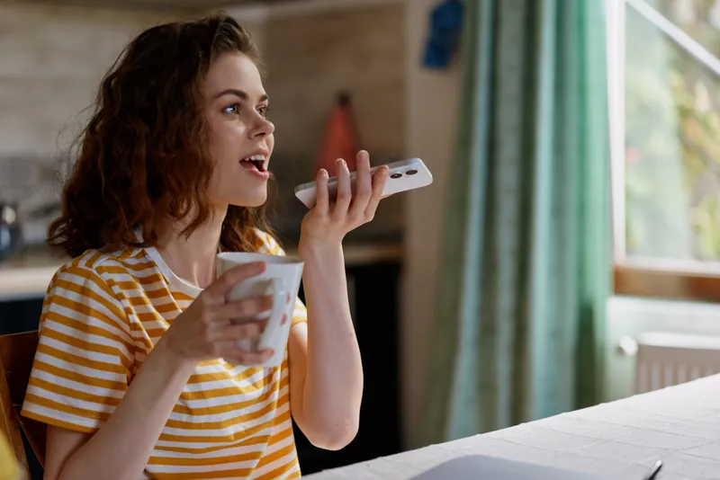 Casual woman at home wearing a striped shirt speaks into a handheld device while sipping coffee by a sunlit window, capturing a relaxed daily routine and warm mood.