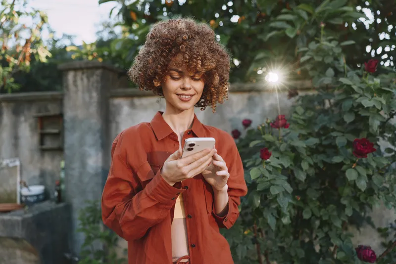 Woman lifestyle portrait outdoors, smiling curly hair, wearing orange jacket, focused on smartphone, relaxed moment in garden with roses, natural light, casual fashion vibe